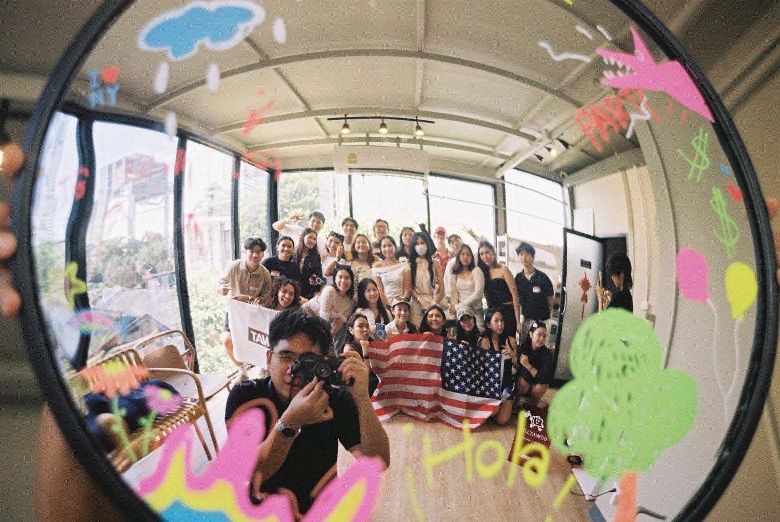 ภาพมุมกว้างผ่านกระจกกลม น้อง ๆ และรุ่นพี่ถือธงอเมริกาถ่ายรูปรวมกันในงาน Dek Work Party