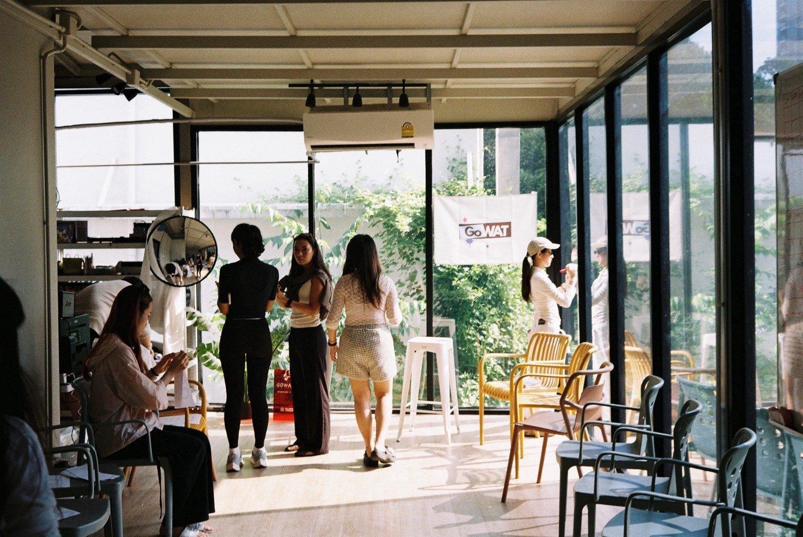 บรรยากาศในคาเฟ่ที่จัดงาน ผู้ร่วมงานยืนคุยกัน มีธง GoWAT แขวนอยู่ด้านหลัง